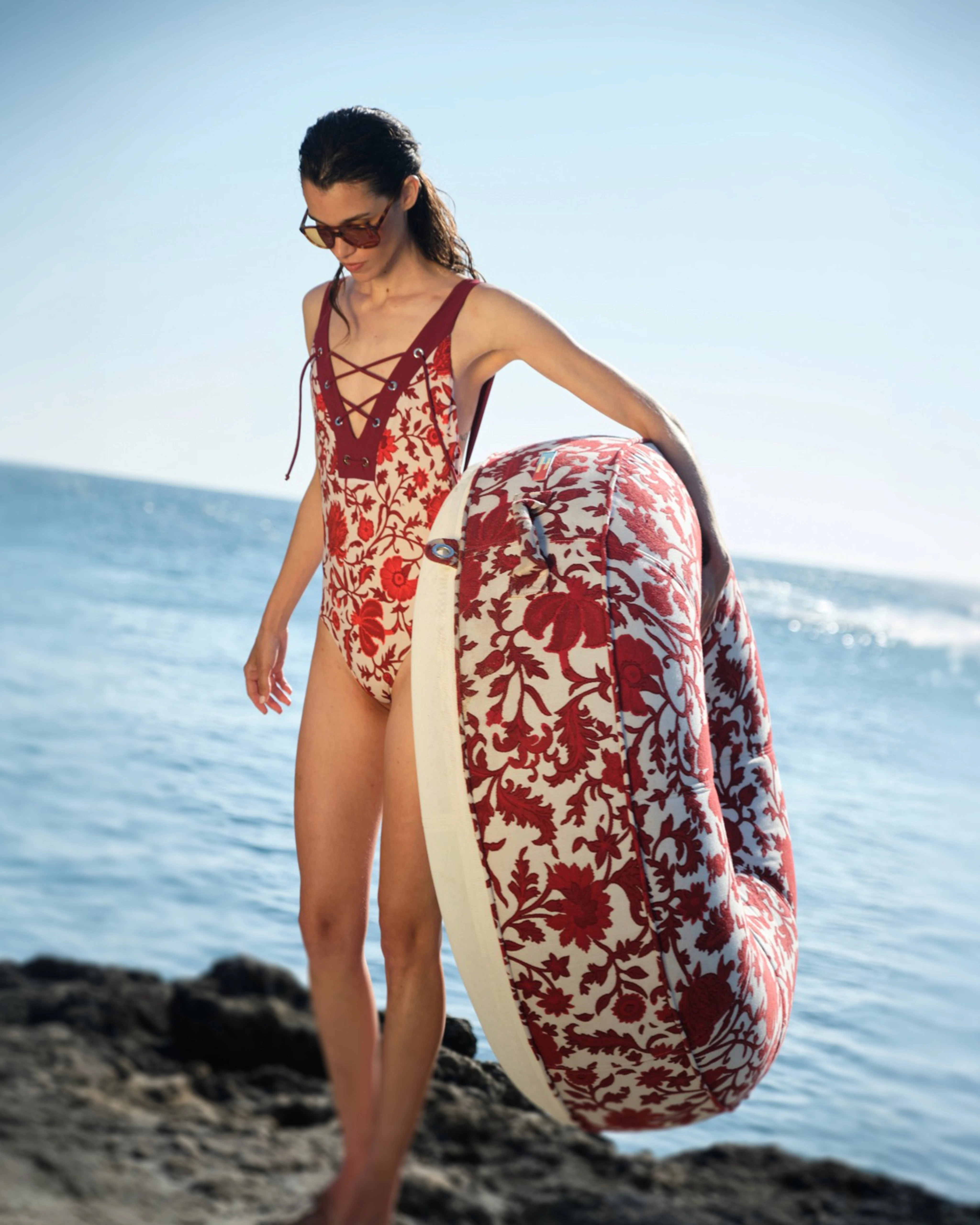 Woman in a matching red floral swimsuit carrying a round luxury pool float with red and cream damask upholstery by Oliver James Lilos — high-end beachside elegance for adults.