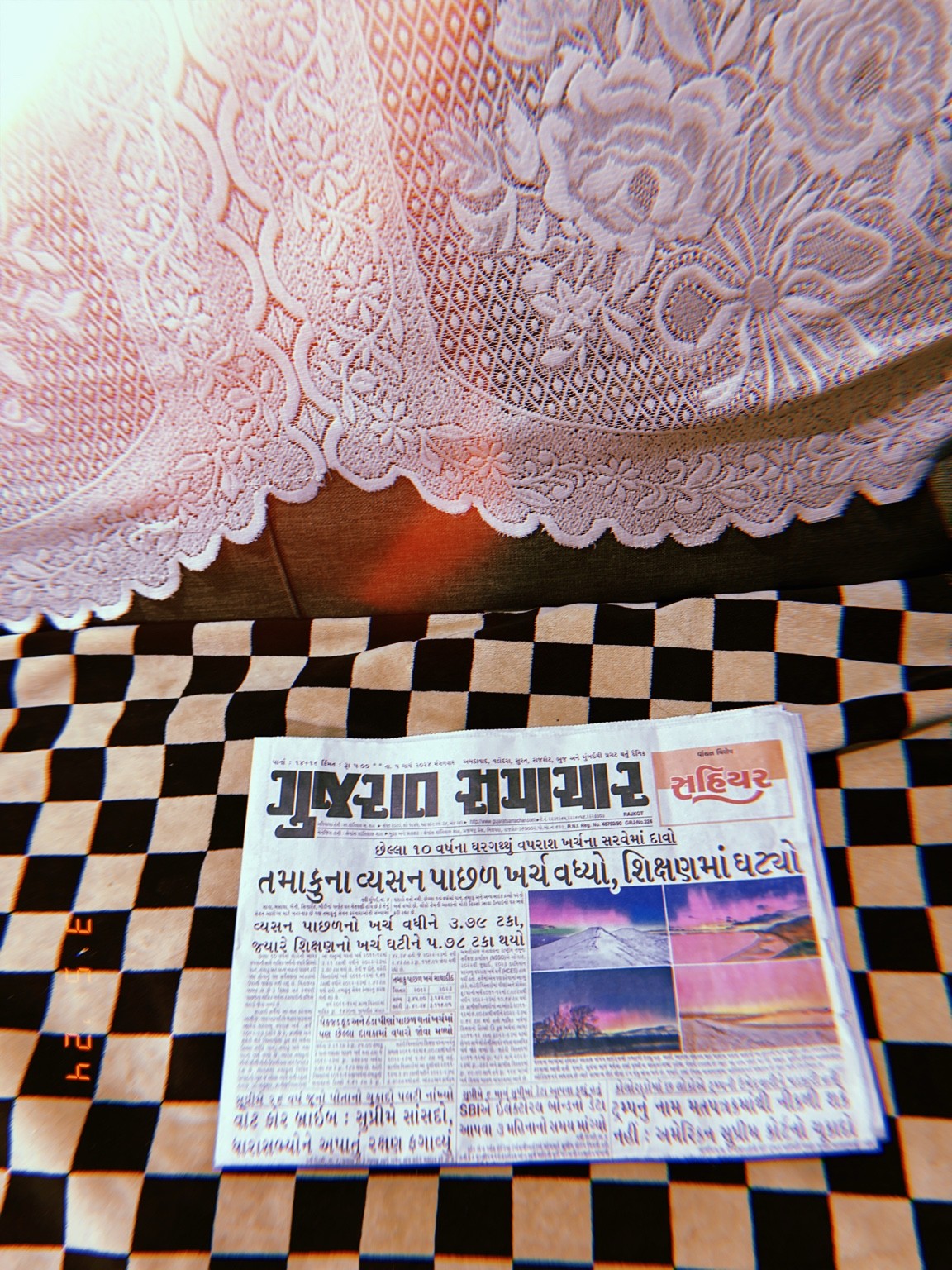 A Gujarati newspaper rests on black and white checkerboard fabric beneath a white floral lace curtain.