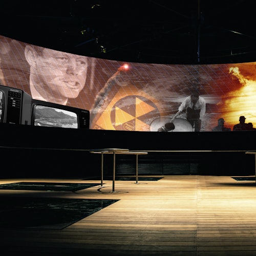 Böjd skärm som visar olika bilder, inklusive en man, en kärnkraftssymbol och en missil, i ett rum med trägolv och tomma bord.