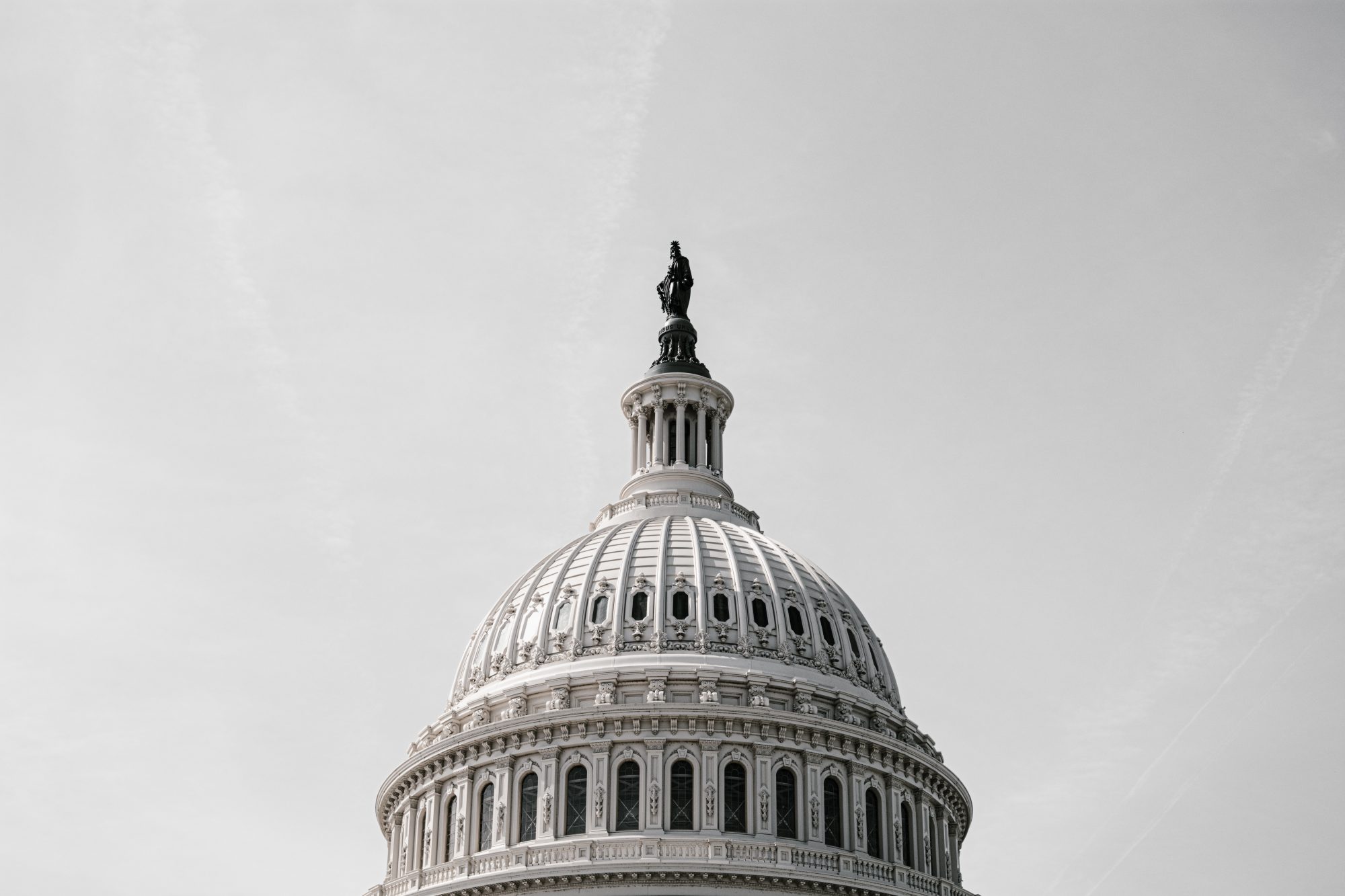 The dome of the U.S. Capitol building against a cloudy sky, focusing on its ornate sculpture atop.