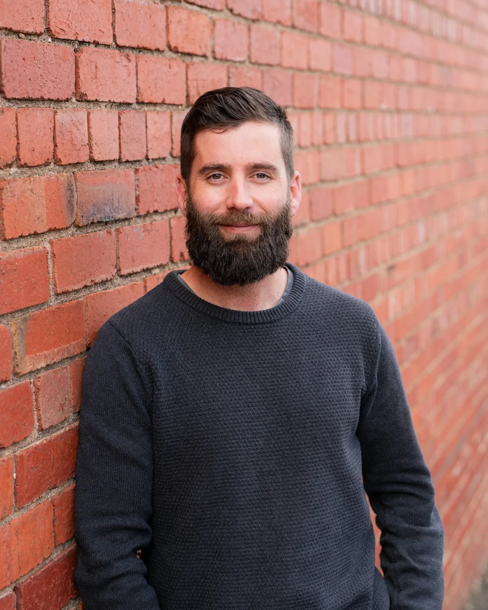 Man with beard smiling against brick wall background.