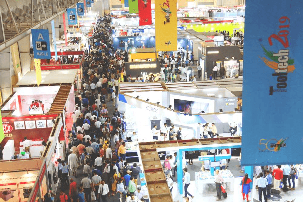 Aerial view of crowded IMTEX FORMING 2026 expo halls at BIEC Bengaluru showing 50k attendees navigating 600+ manufacturing technology booths across 46,000 sqm, live demos with laser sparks and glowing hydroforming presses visible from above