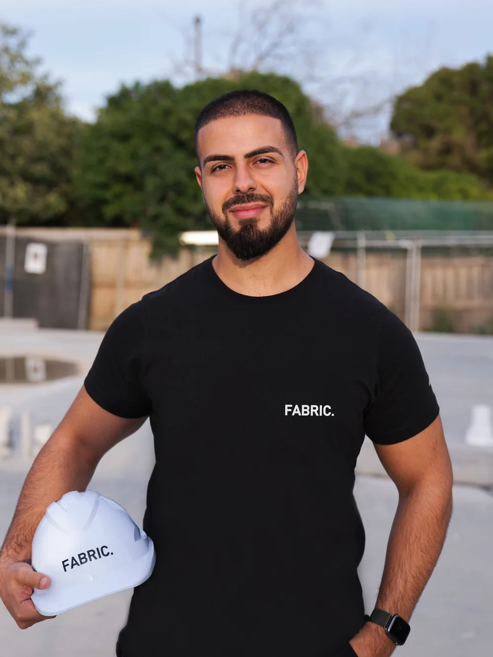 Man holding a white hard hat outdoors wearing a black FABRIC t-shirt.