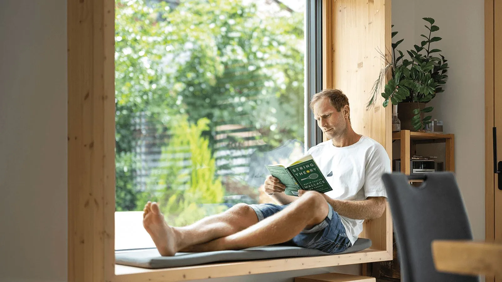 Ein Mann liest ein Buch namens "String Theory" auf einer Fensterbank mit Blick ins Grüne.