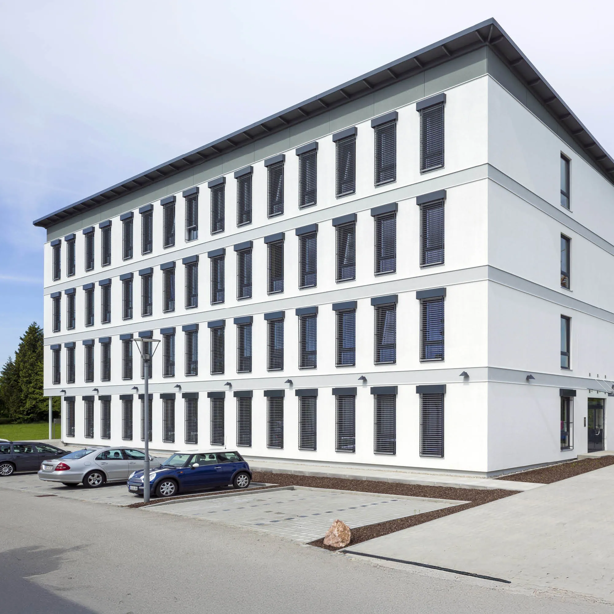 Modernes Bürogebäude mit weißen Fassaden, vielen graugrünen Fenstern und Jalousien, Parkplätzen und einem Baum.