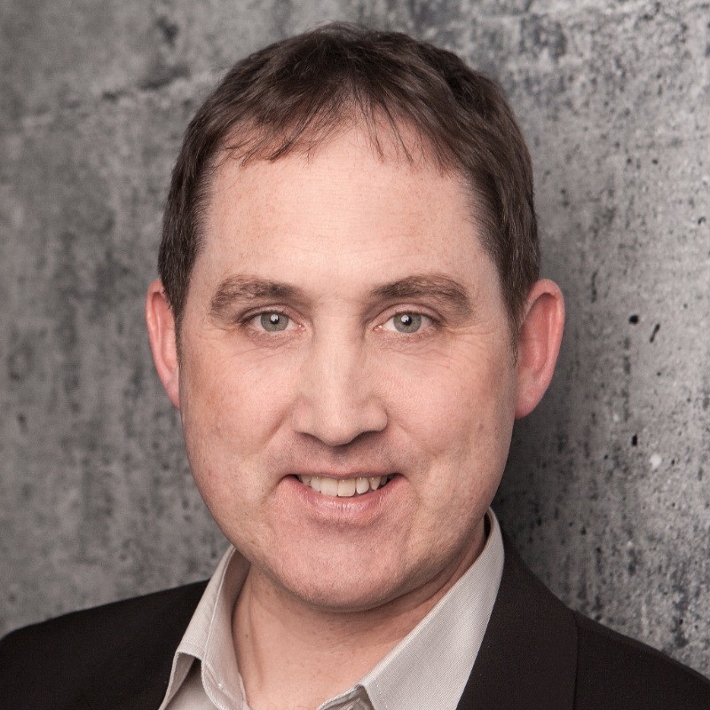 Smiling young woman with curly hair sitting at a laptop, in front of a teal wall.