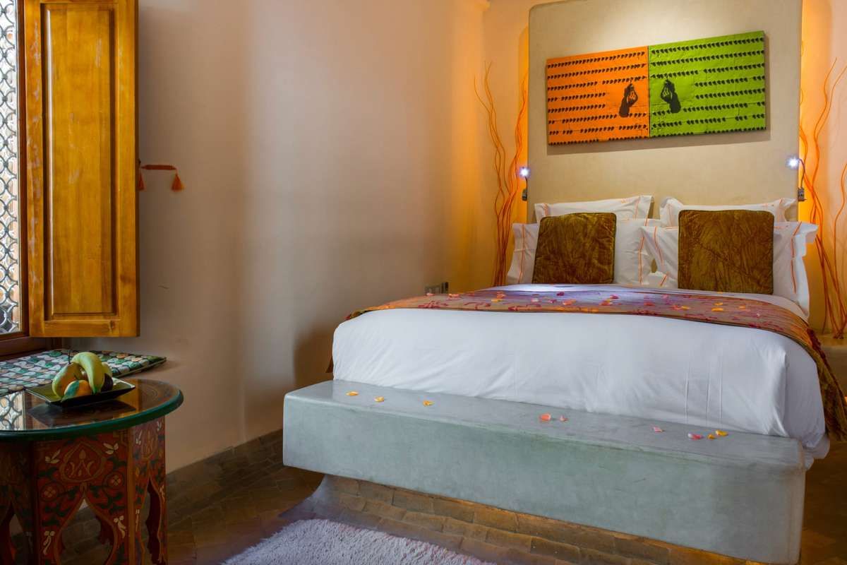 A minimalist bedroom with a white bed and patterned pillows sits beneath a large orange and green mural on a plain wall.
