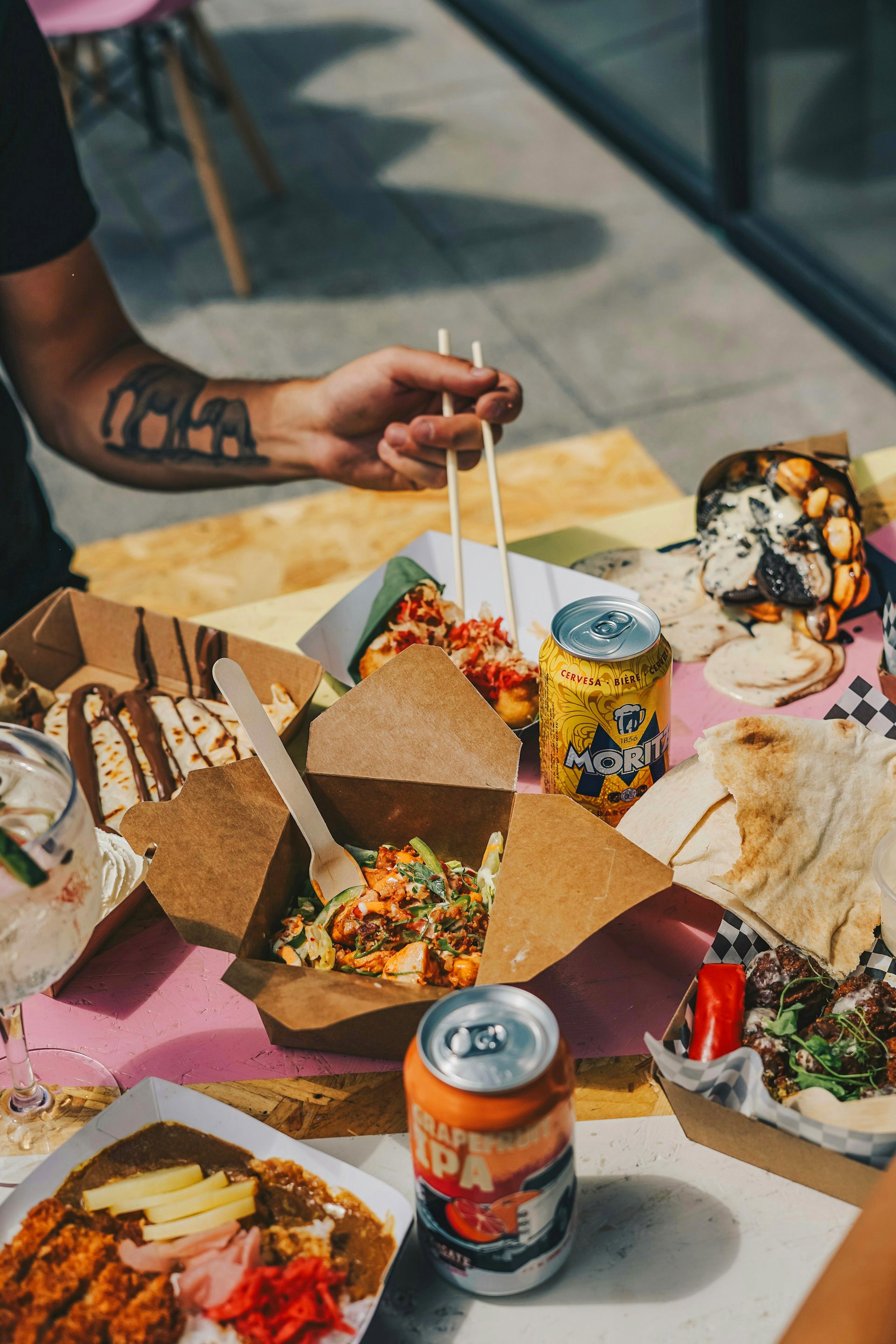 A person with an elephant arm tattoo uses chopsticks to eat from a takeout box amidst a spread of street food, desserts, and beer.