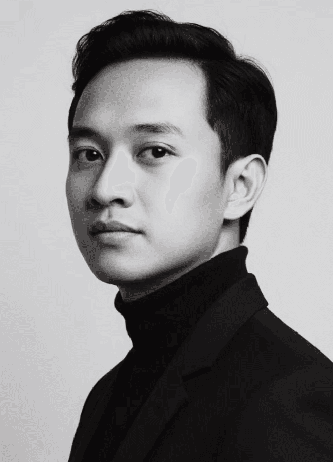 Man sitting in a dimly lit room, wearing a beige shirt and looking contemplative.
