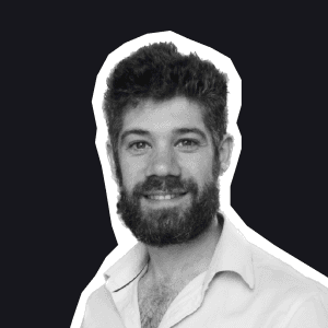 A smiling man with a neatly trimmed beard and dark hair, wearing a light shirt, against a dark background representing a.
