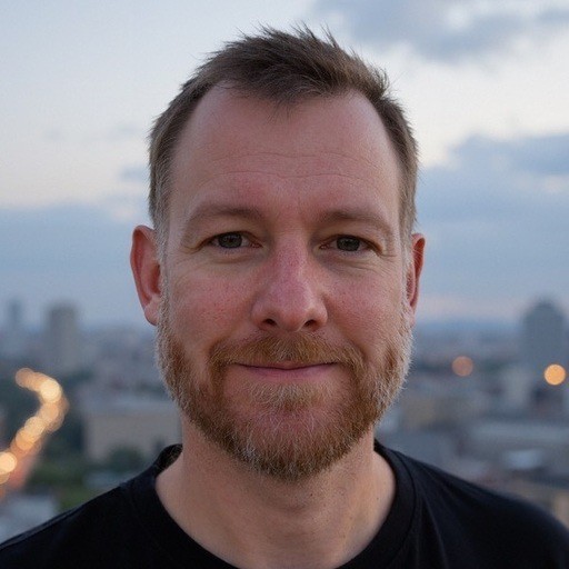 Young man in an orange hoodie and glasses at a desk with dual monitors in a dark office 