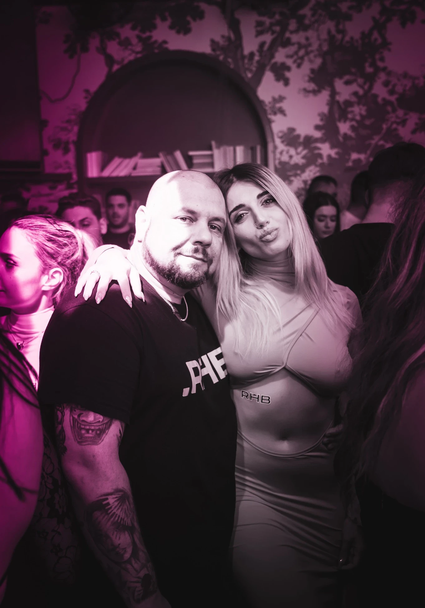 Provocateur with a beard and tattoos poses with his arm around a woman. The woman, wearing a cutout dress with an "RHB" sticker, has her arm on his shoulder and is making a kissy face for the camera.