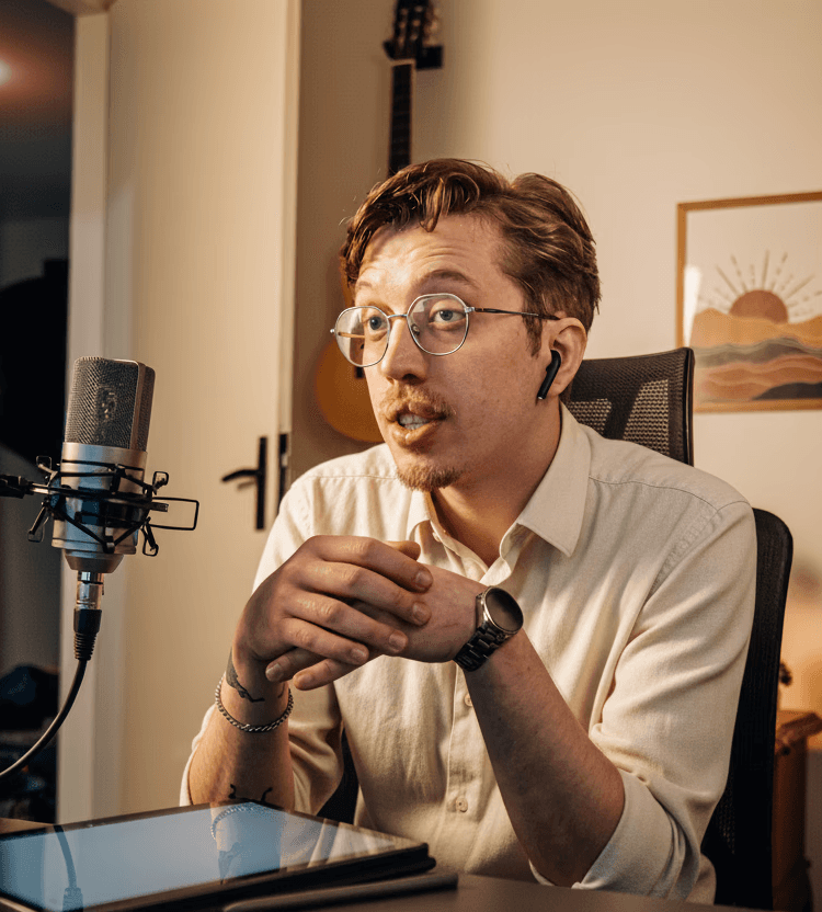 Homem de óculos e fone de ouvido sem fio, usando camisa clara e falando em um microfone profissional. Ele está sentado em uma mesa com um tablet, em um escritório doméstico com violão, quadros de arte e um estetoscópio pendurado ao fundo.