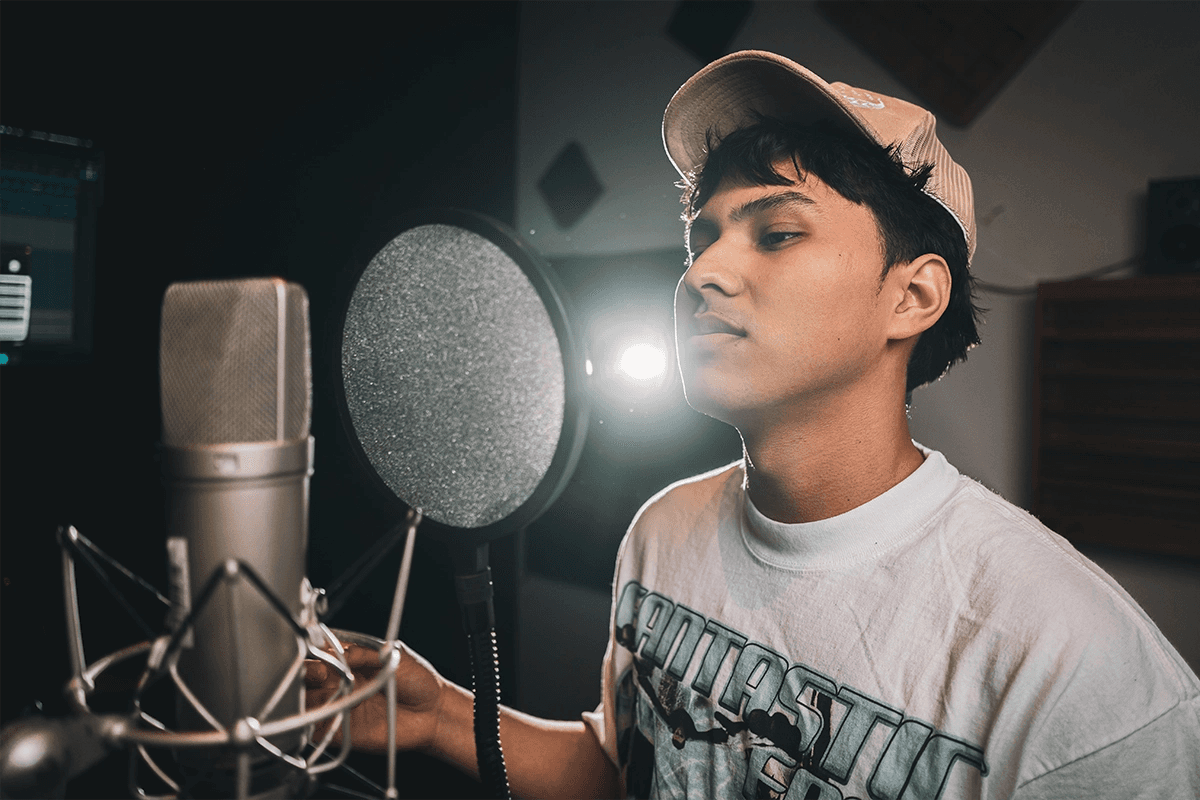 A young man wearing a cap and graphic T-shirt is standing in front of a large microphone in a recording studio, intently focused as he performs into a pop filter with acoustic panels visible in the background.