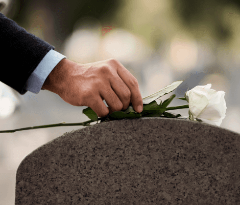 A close-up of a hand gently placing a white rose on a gravestone. The hand is dressed in a black suit sleeve, and the scene conveys a sense of remembrance and respect. The background is softly blurred, focusing on the act of honoring a loved one.