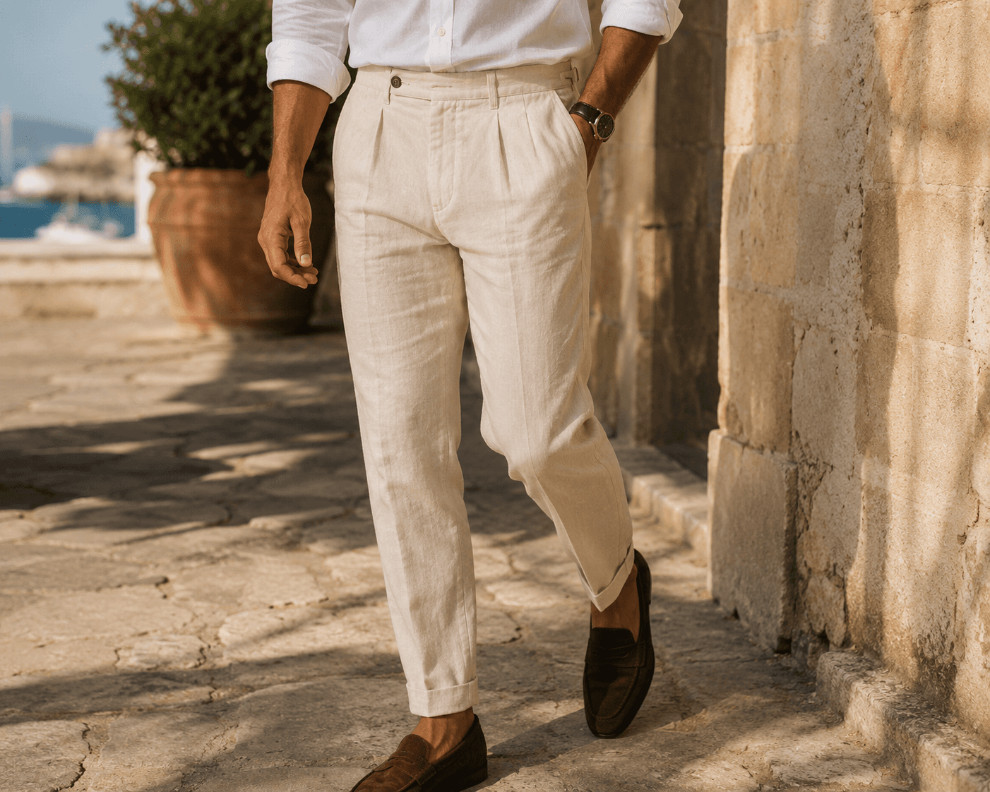 A mid-section shot of a man walking outdoors along a sunny, stone-paved coastal path in classic resort wear. He is dressed in a crisp white linen shirt with rolled-up sleeves, tailored off-white pleated linen trousers with cuffed hems, and dark brown suede penny loafers. He wears a classic watch with a leather strap on his wrist, and a large terracotta potted plant is visible in the soft-focus background.