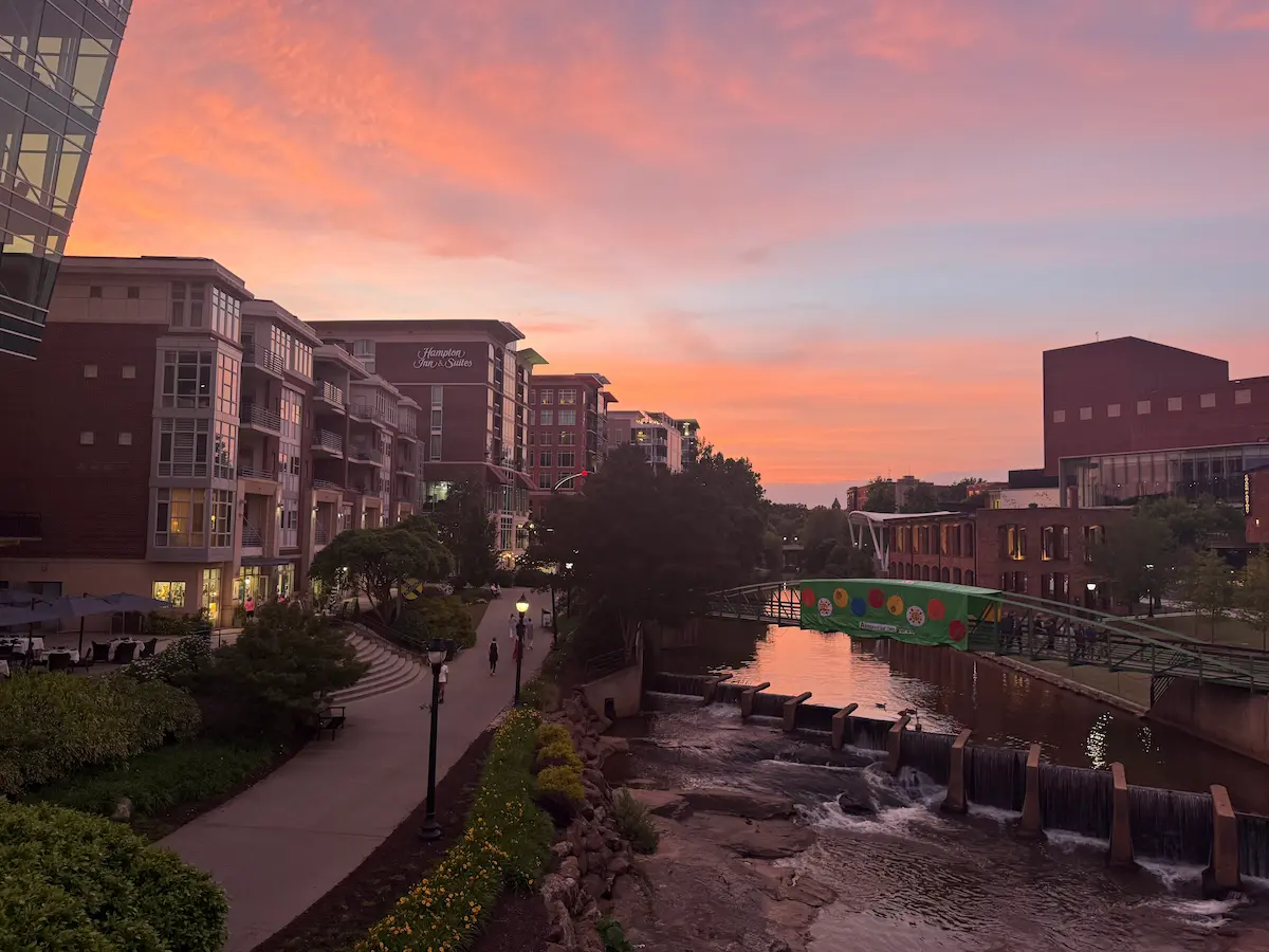 Sunset over the Reedy River in downtown Greenville, South Carolina, with pink and orange clouds reflecting above the riverwalk, bridge, and city buildings.
