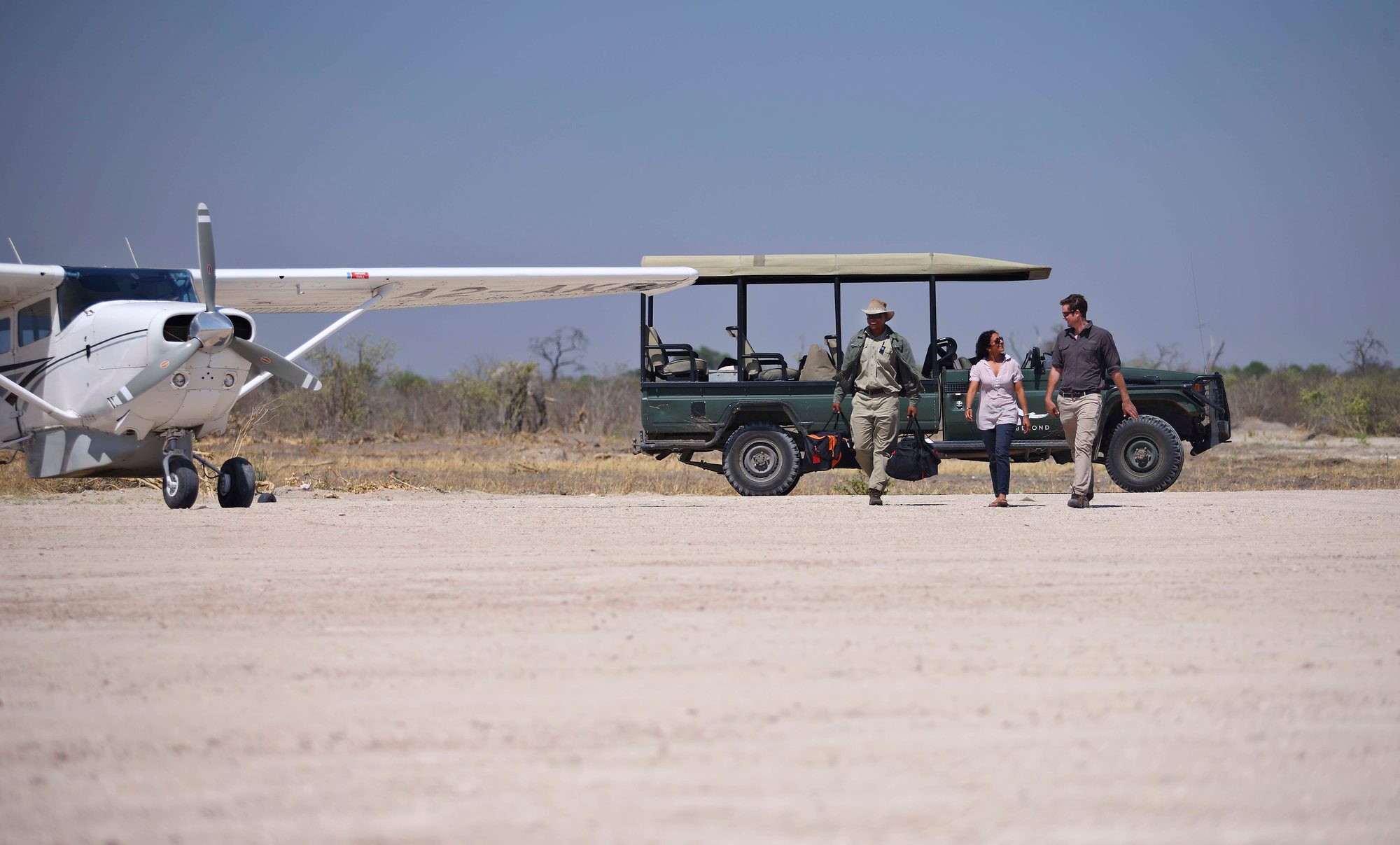 A guide carries bags as two guests walk across a sandy airstrip past an open safari vehicle, with a light aircraft parked nearby in Botswana.