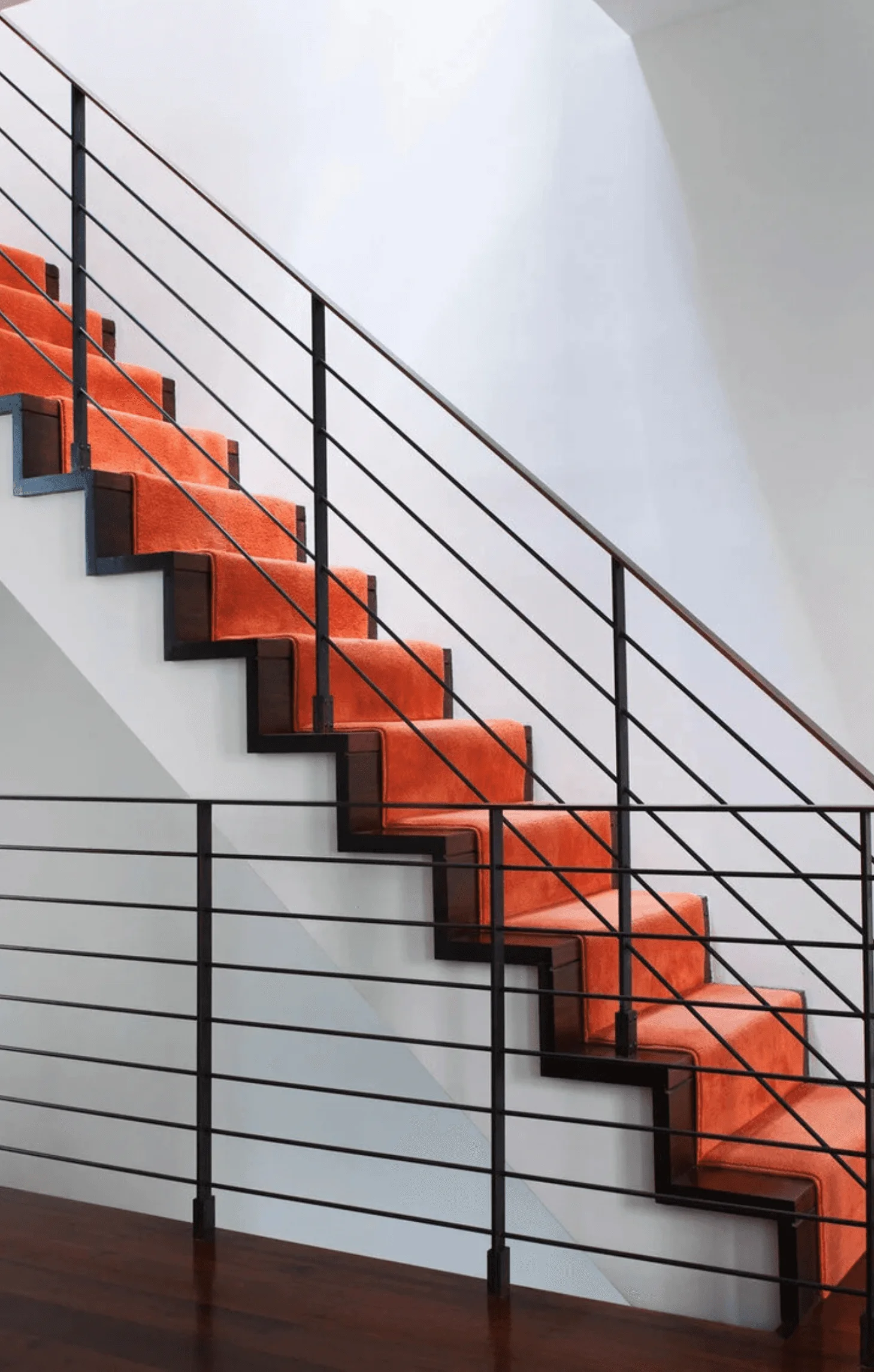 An orange stair runner climbs a black iron staircase against white walls
