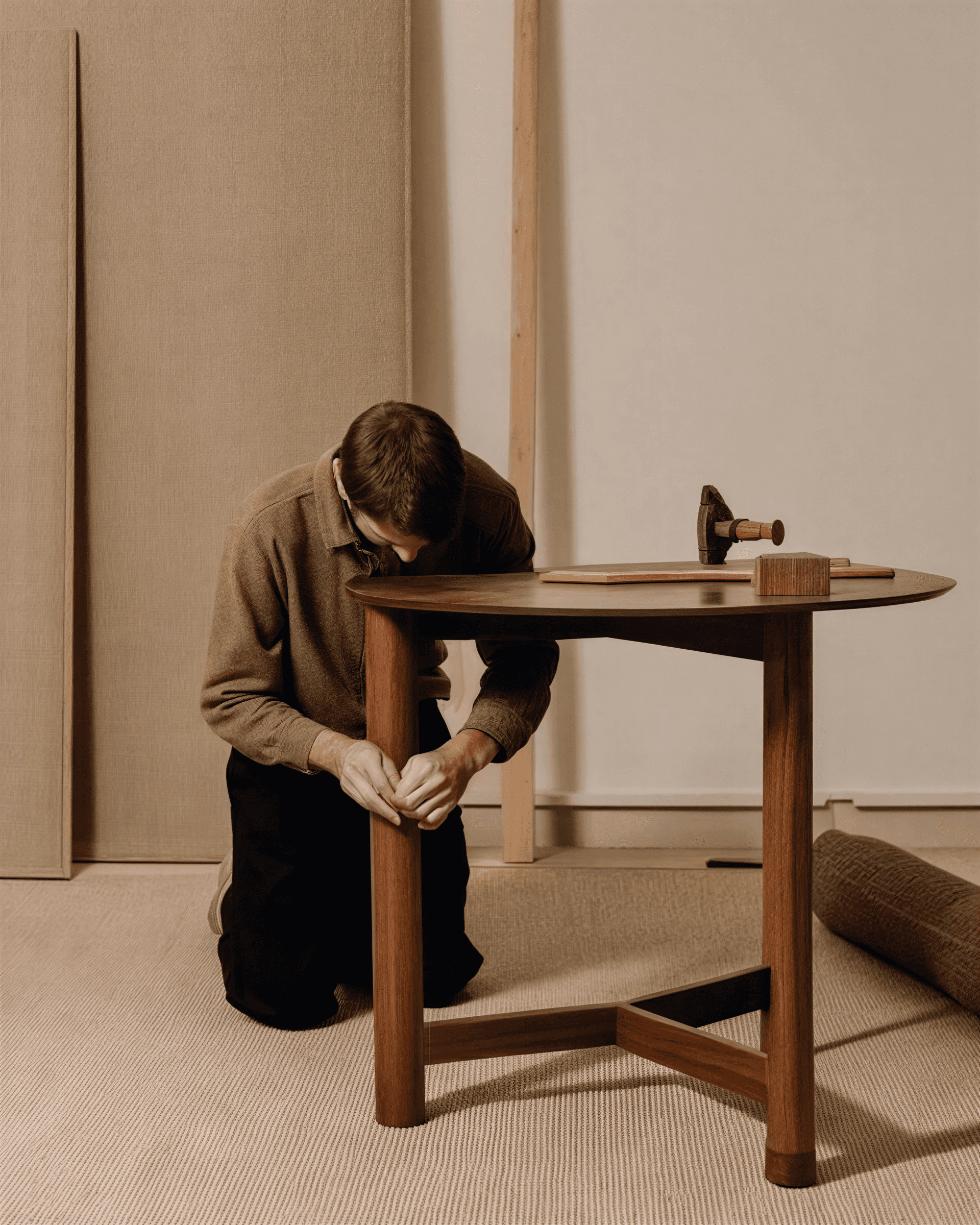Man kneeling on a beige carpet, assembling a wooden table with tools nearby in a minimalist workshop setting.
