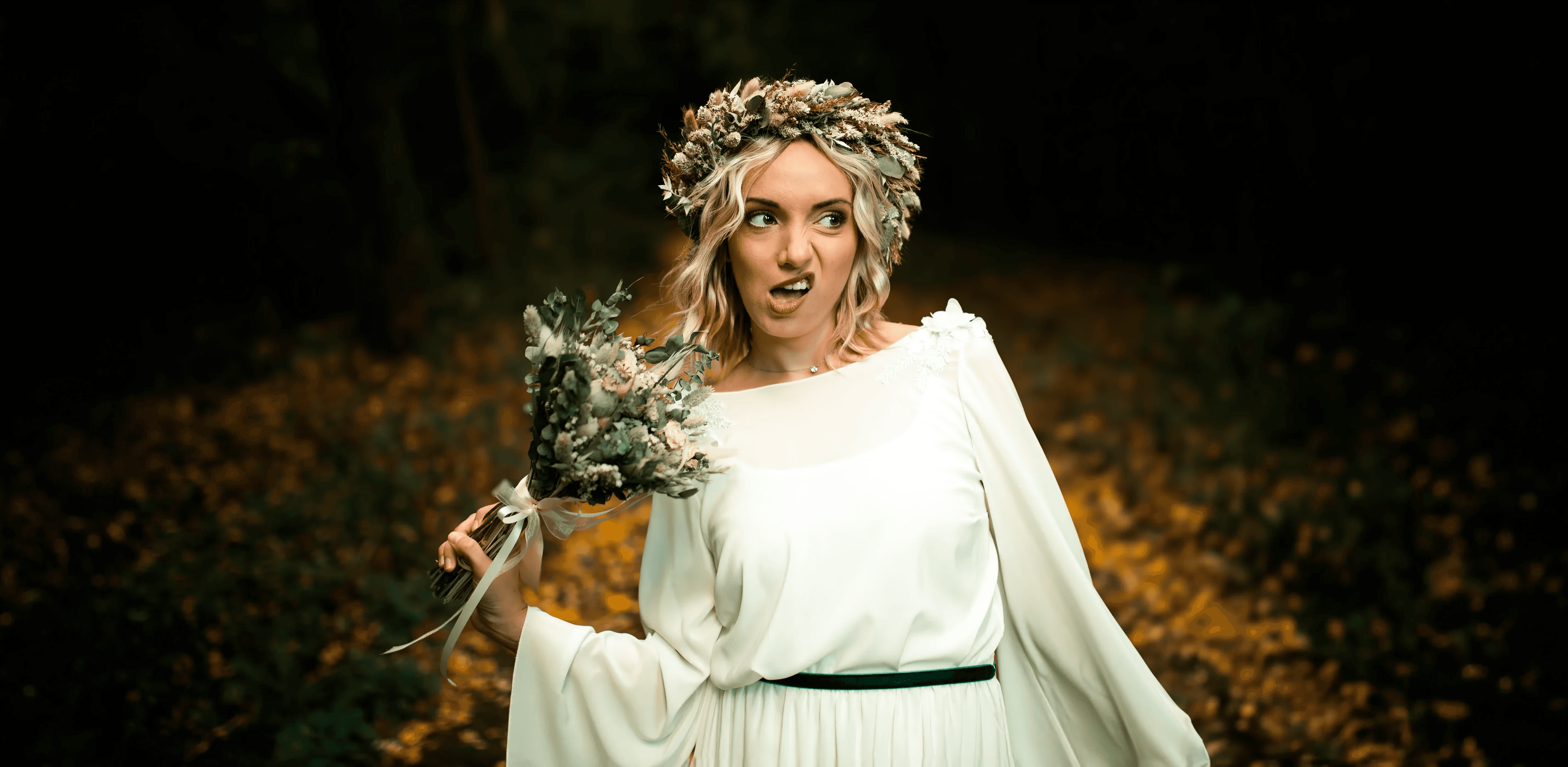 Játékos menyasszony hullámos szőke hajjal, szárított virágkoszorúval és egyszerű fehér hosszú ujjú ruhában zöld övvel, boho csokrot tartva, Tóvik vendégház esküvői fotózás