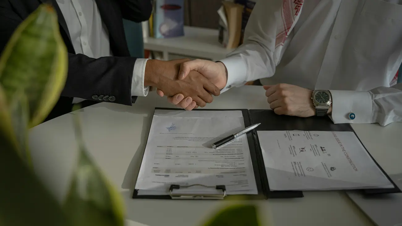 Two business representatives shaking hands over a signed contract on a desk, illustrating a sponsorship agreement between a sports club and a commercial partner.