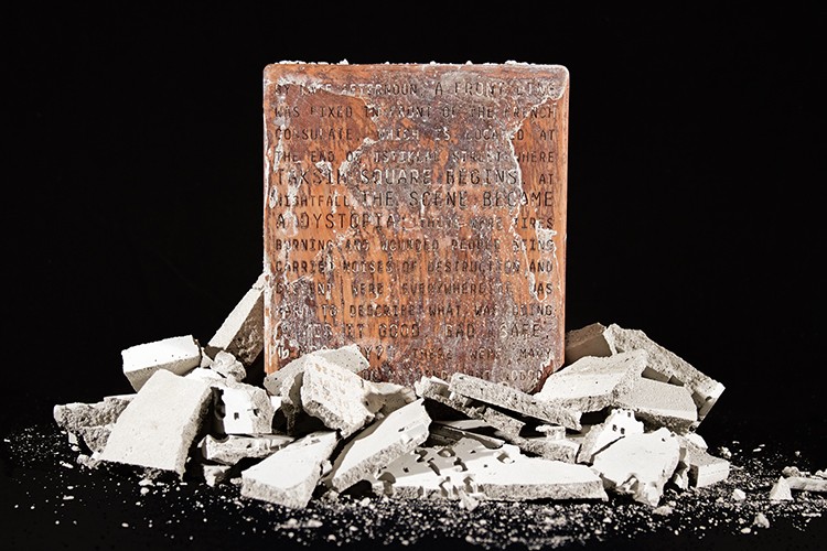 A rectangular, light brown stone slab with inscribed text sits atop a pile of broken white stone pieces against a black background. The slab’s surface shows signs of weathering and wear, with visible cracks and uneven texture. The broken pieces are scattered around the base of the slab, some resting directly on its surface. The contrast between the smooth, weathered stone and the fragmented, broken pieces emphasizes the slab’s central position and the text it bears.