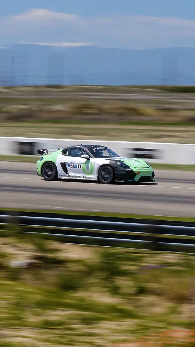 A green and white Porsche race car speeds along a track with mountains in the background under a clear sky.