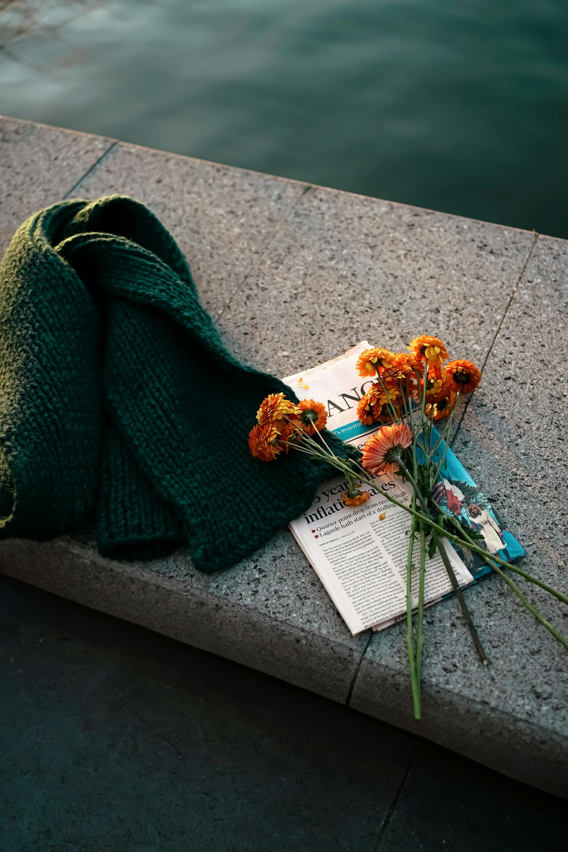 A dark green knitted scarf, a bouquet of orange gerbera daisies, and a newspaper lie on a granite ledge next to a body of water.