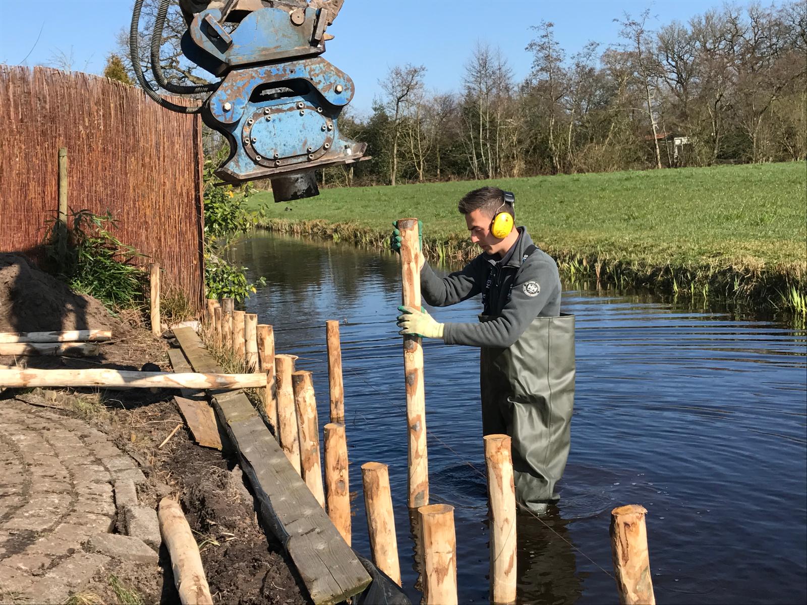 Hovenier plaatst houten palen in oever met kraan voor nieuwe tuinbeschoeiing