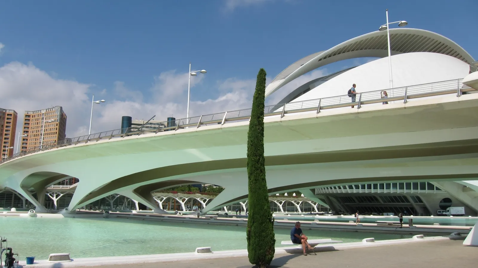 A modern, futuristic bridge with sleek arches spans across a serene, turquoise water body, set against a backdrop of a clear blue sky and tall, contemporary buildings in the distance.