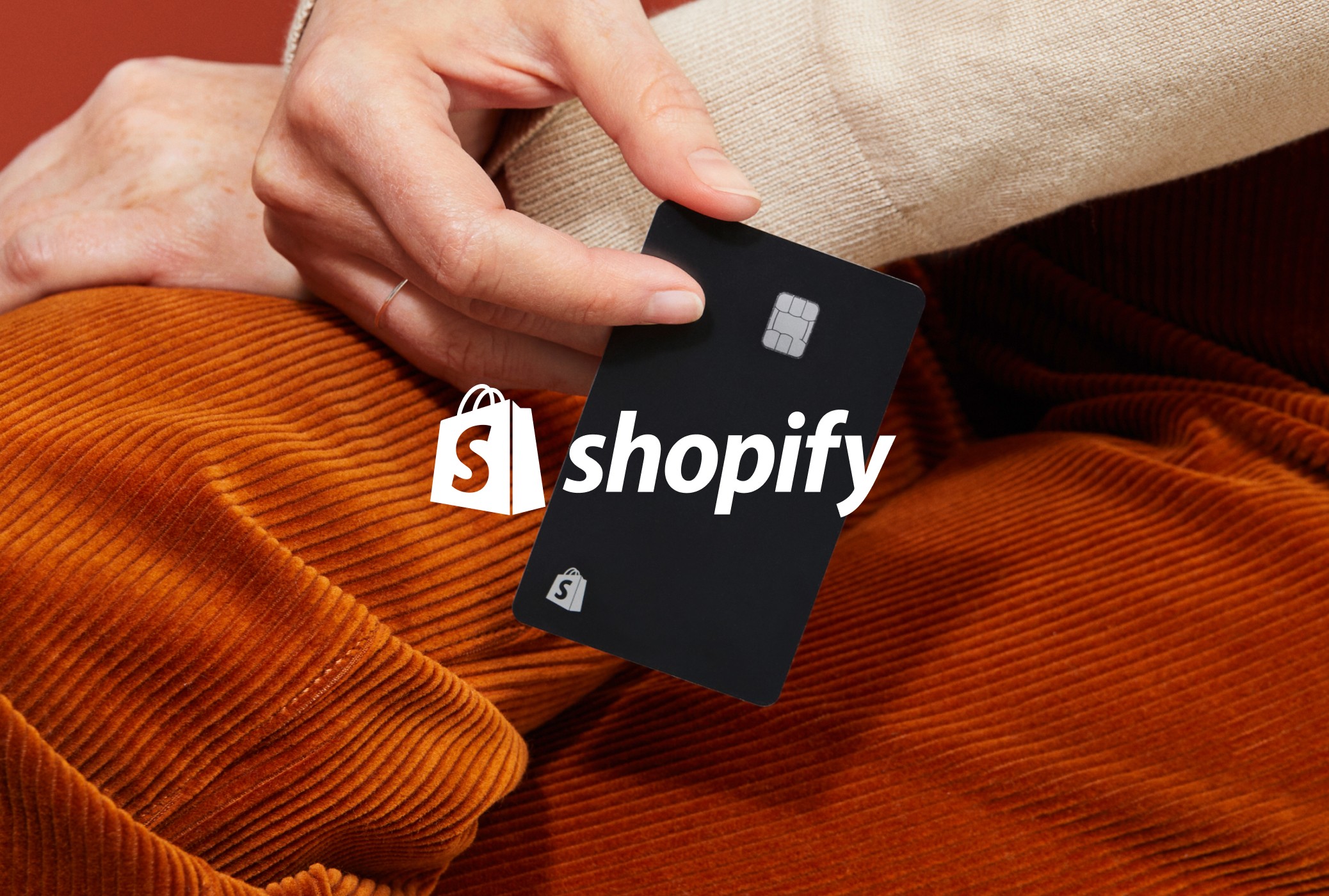 A hand holding a black Shopify card, resting on orange corduroy beside the Shopify logo