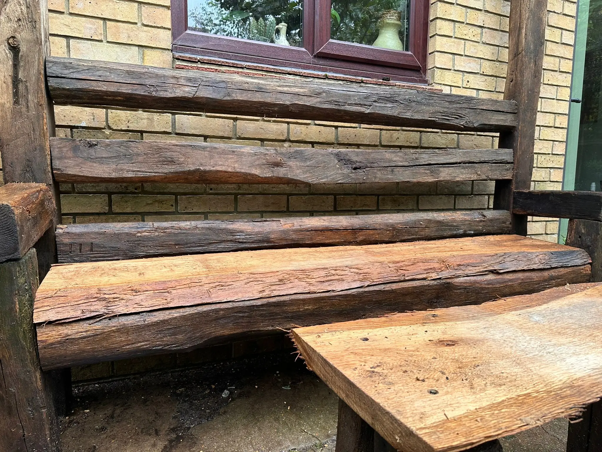 A rustic wooden bench made from logs, positioned beside a wooden table against a brick wall.