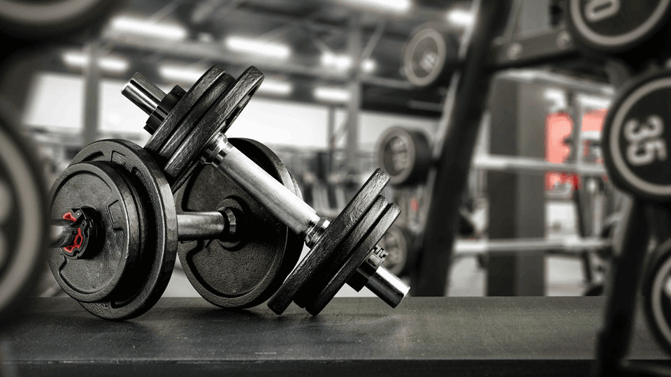 A close-up of a weightlifting setup with dumbbells in a gym, featuring a blurred background of exercise equipment.