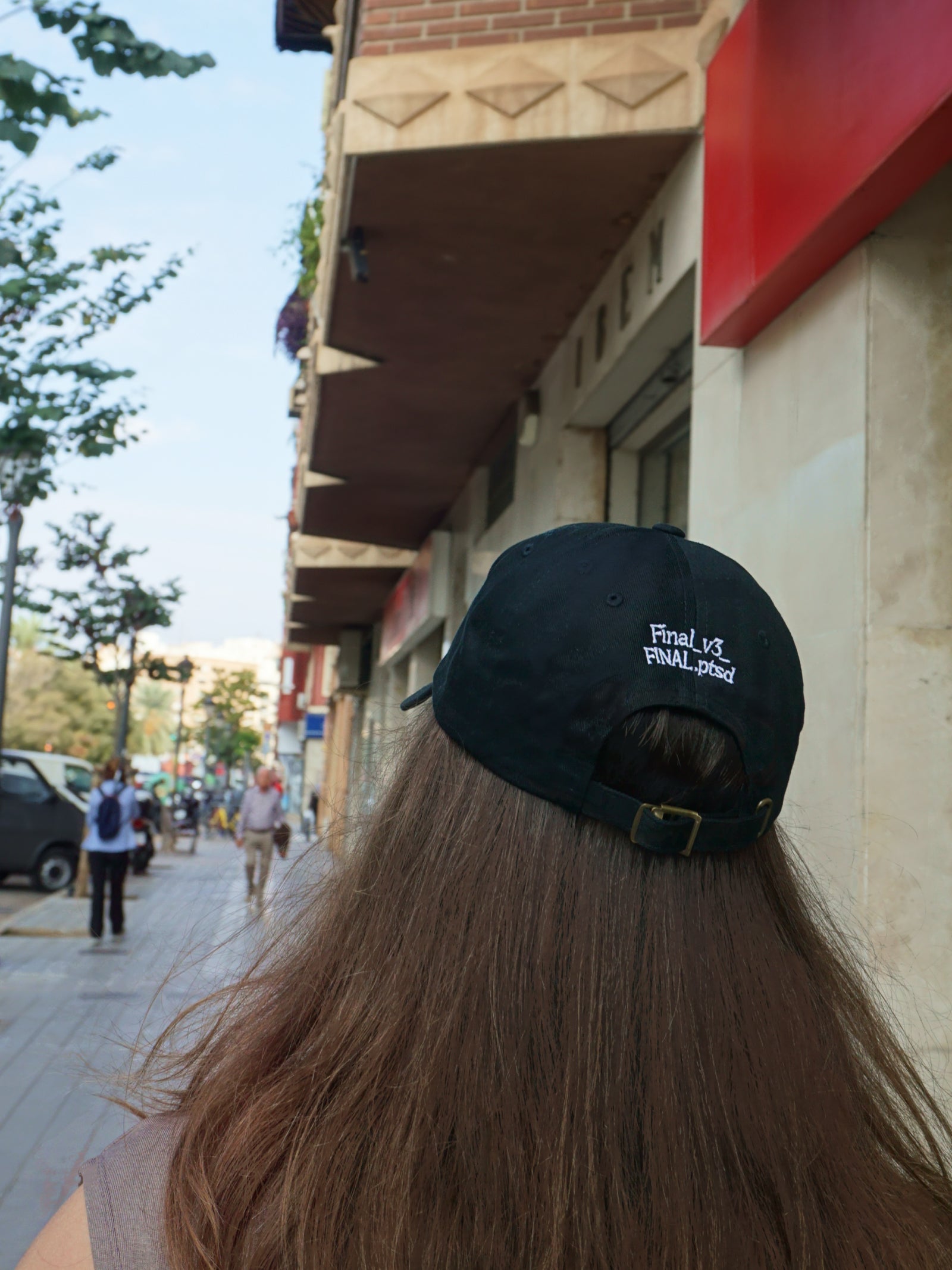 Person wearing a black cap with text, walking on a street with buildings and people in the background.