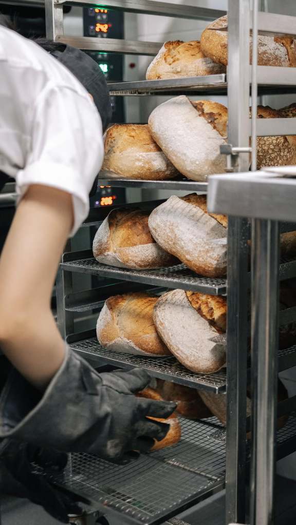 A baker wearing gloves retrieves fresh sourdough loaves from a metal rack in a bakery setting, showcasing an array of artisanal breads.