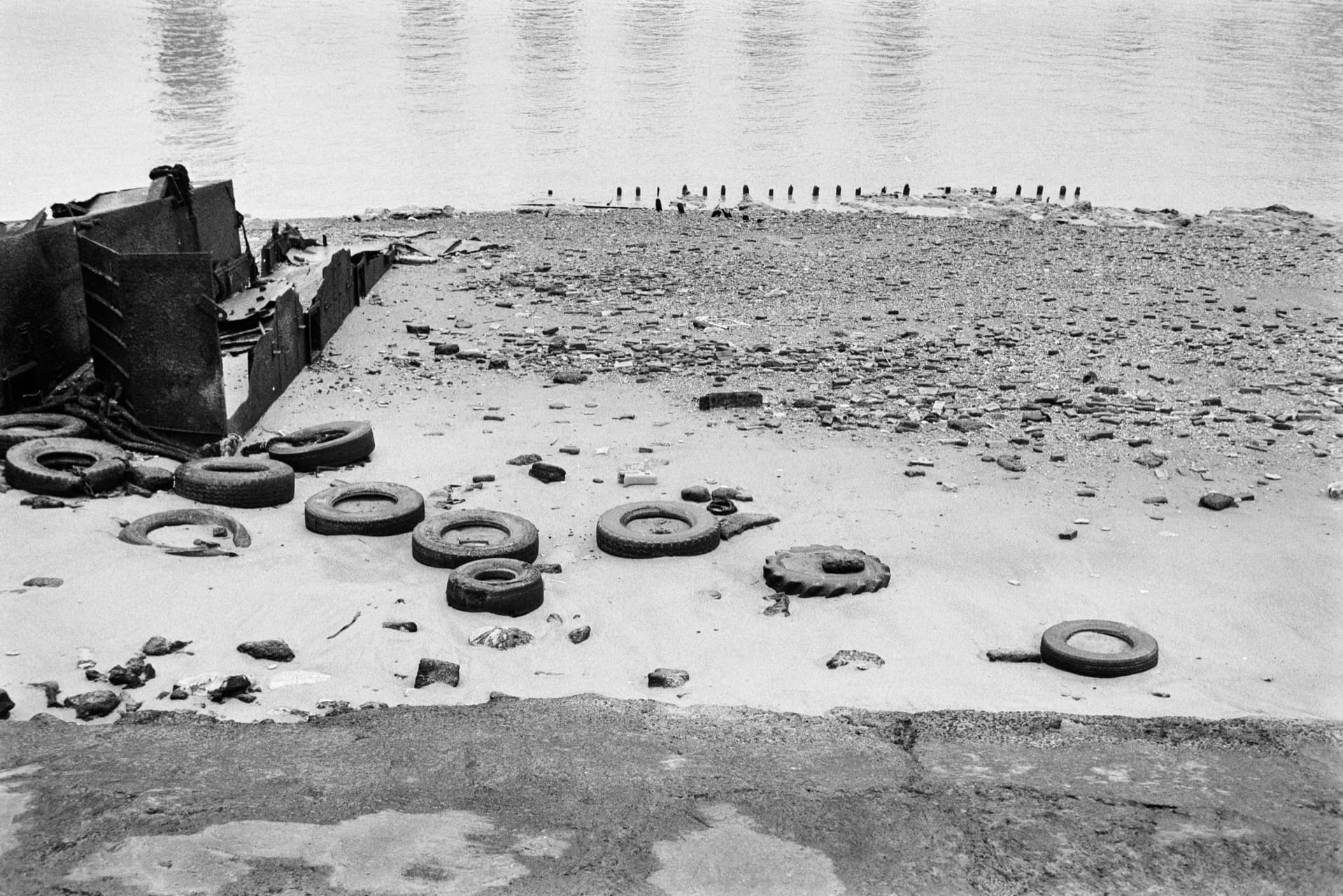 Thames foreshore at North Greenwich with scattered tyres and debris, collapsed metal structure at left, wooden posts extending along shoreline, industrial river visible beyond