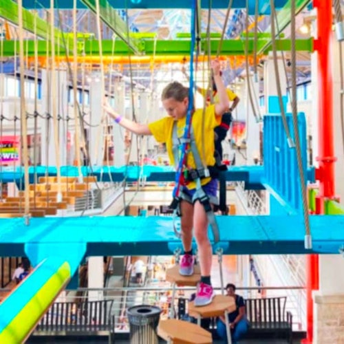 A child in a yellow shirt navigates a colorful indoor ropes course, wearing safety gear, with people in the background.