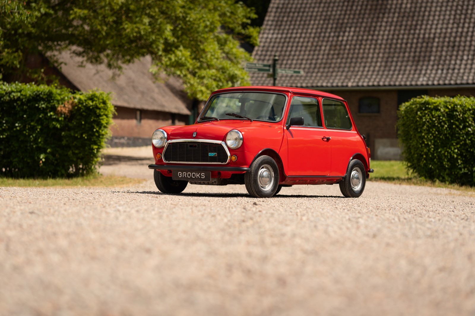 A red car parked on a gravel driveway surrounded by green hedges and a rustic building in the background.