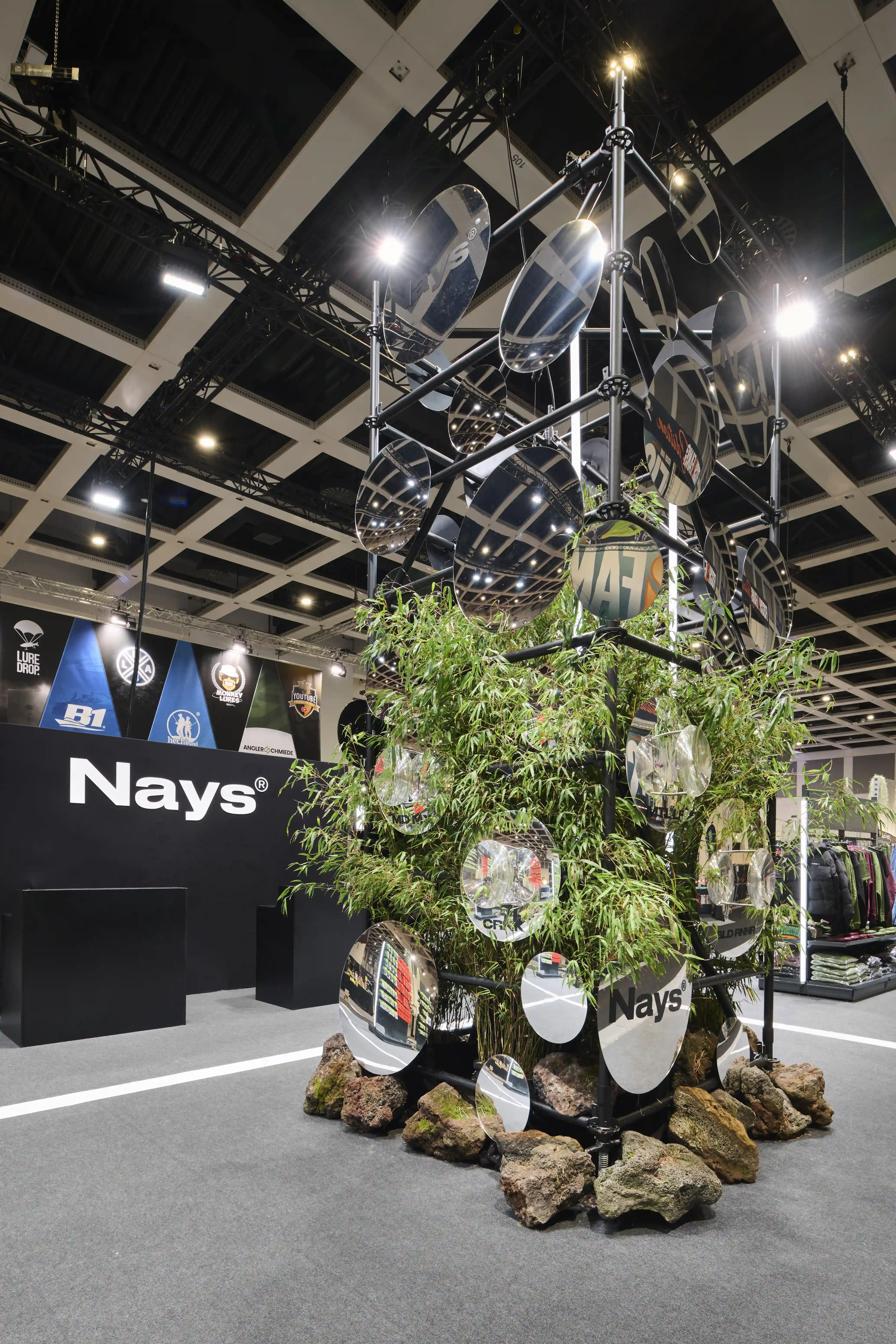 Tower of round mirrors and bamboo plants on lava rocks next to the black Nays logo wall in the exhibition hall.