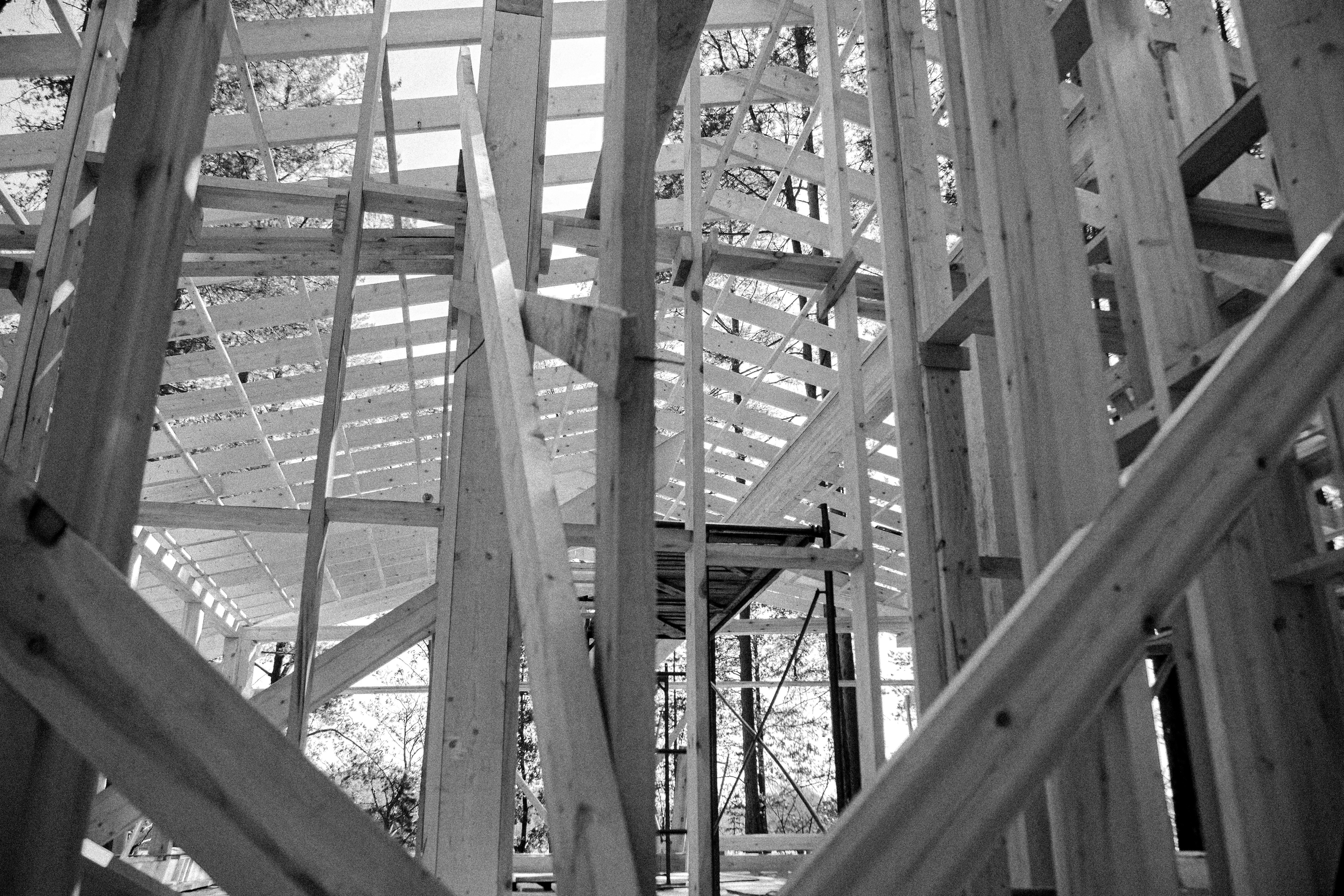 Modern architectural workspace with large floor-to-ceiling windows, surrounded by trees, and desks filled with design materials and books. Two blurred figures are walking through the space.