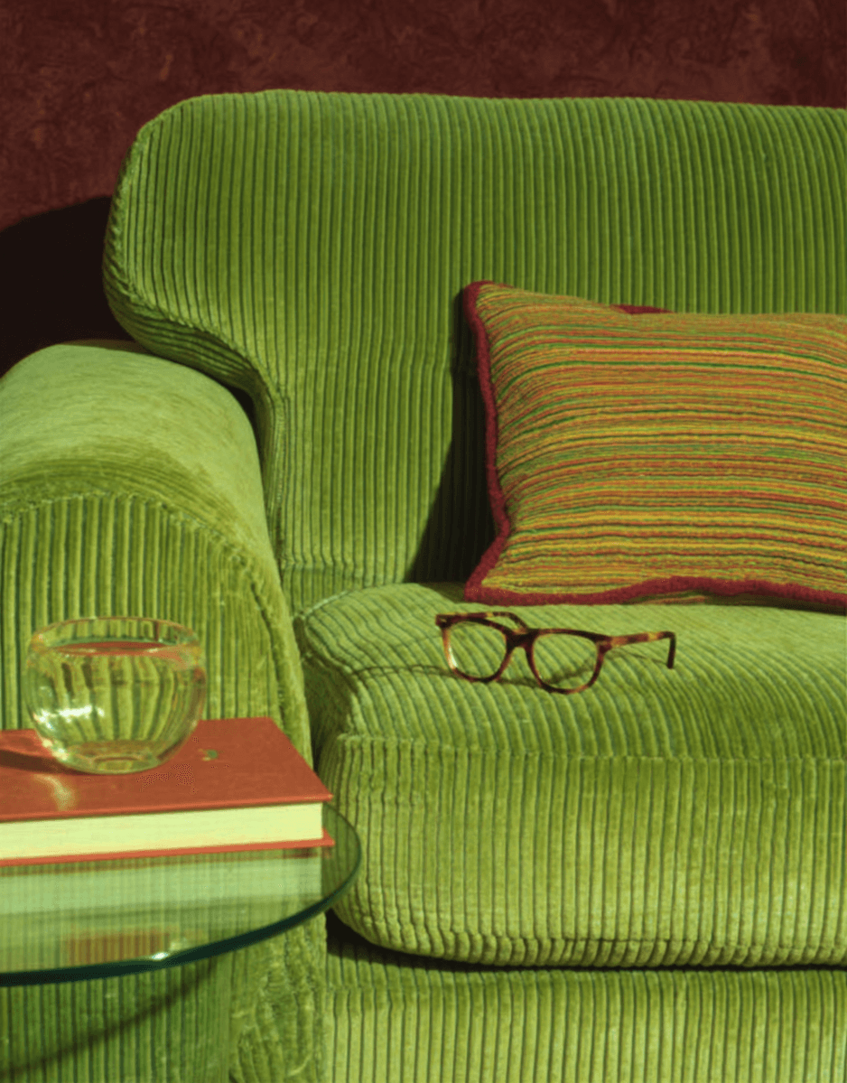 Green corduroy sofa with patterned cushion, eyeglasses on the seat, and a glass of water on a book beside the couch.