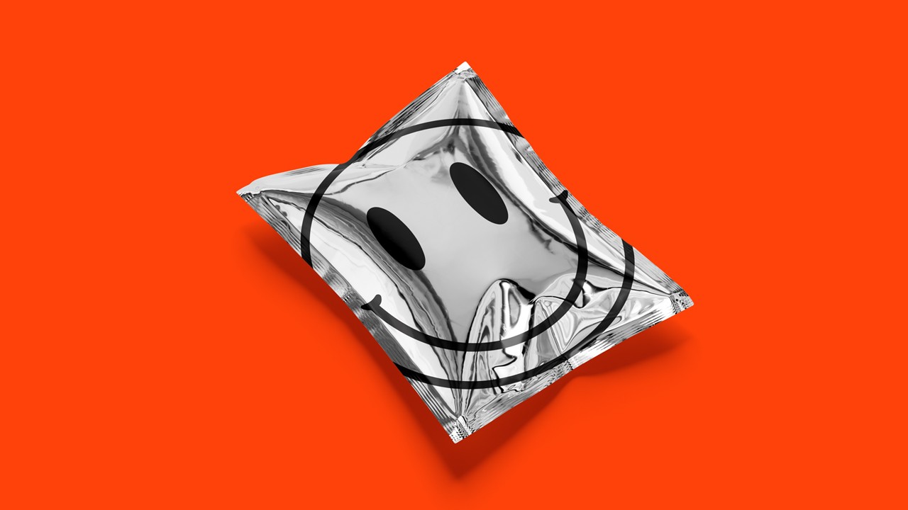 Shiny metallic silver foil pouch lying flat, printed with a large black outline of a smiley face, all set against a bright orange backdrop.