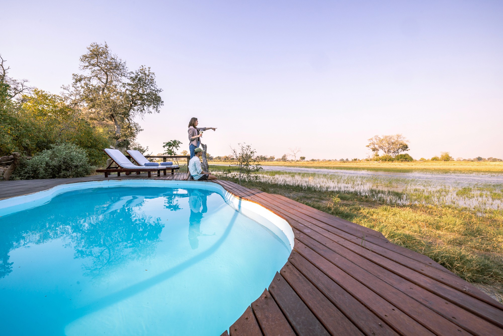 Kujwana Delta Ride, Okavango Delta Botswana. Mjuk loungesoffa och soffbord placerat på pooldäck där två personer njuter av utsikten. Pool med utsikt över deltats vackra landskap.