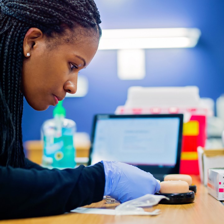 A woman wearing gloves focuses intently on a medical training model. A laptop and medical supplies are in the blurred background, suggesting a clinical setting.