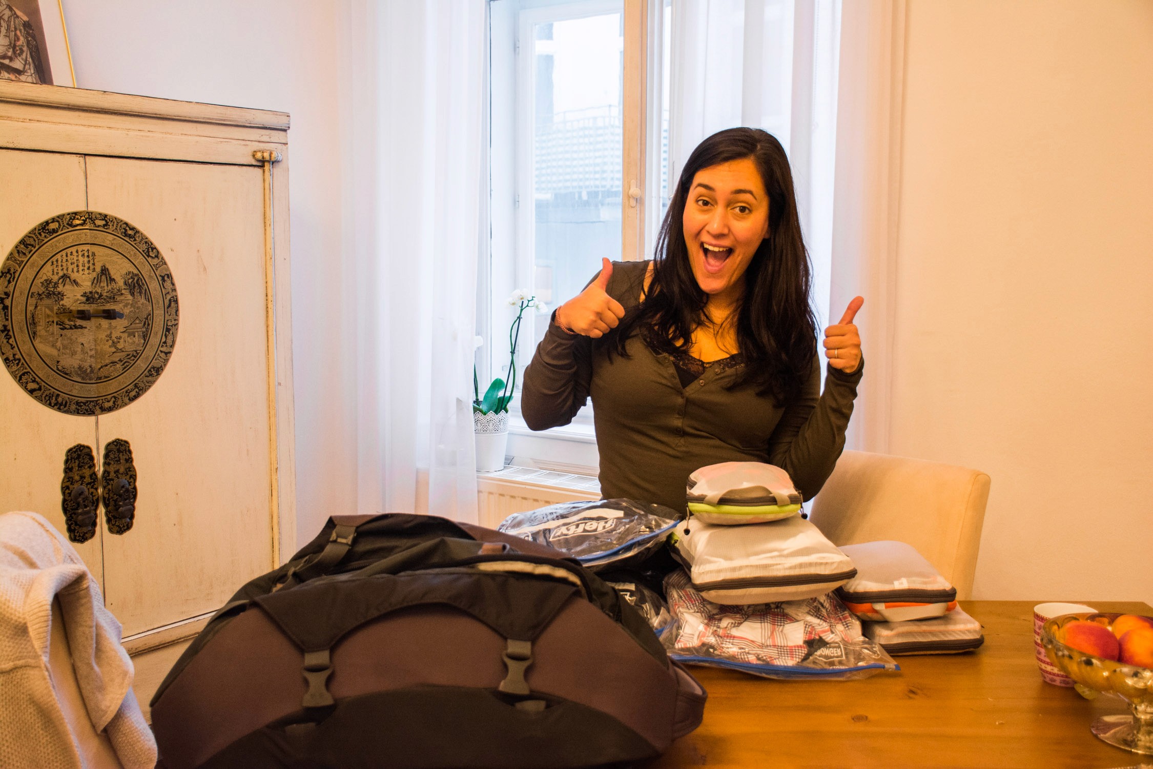 A smiling woman with long dark hair sits at a table covered in travel gear and bags next to a vintage wooden cabinet.