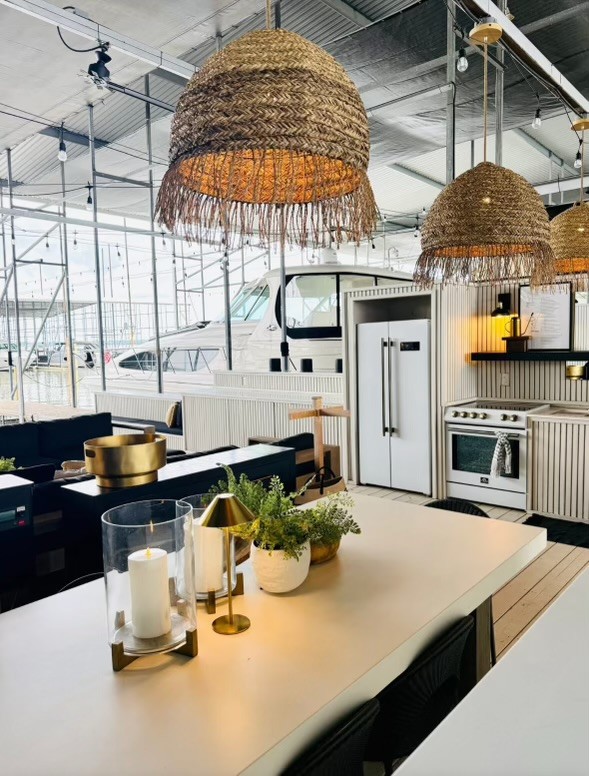 A modern, nautical-themed kitchen with large wicker pendant lights, sleek white cabinetry, gold accents, and a view of docked boats outside.