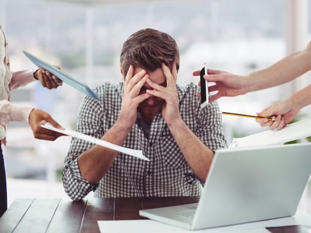 A man sitting at a desk, looking stressed with his hands on his head, possibly overwhelmed by work.