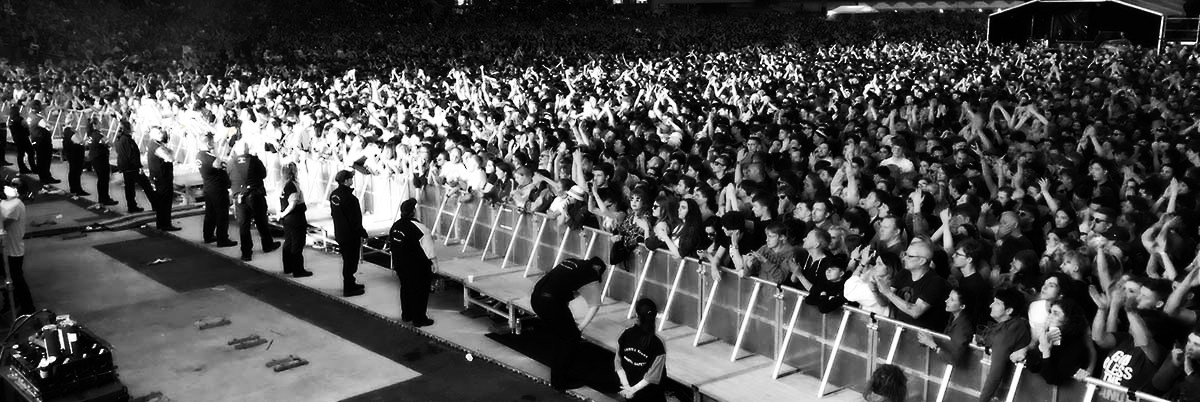 Black and white photo of a large concert hall with view from the side of stage with security staff in the pit and large crowd cheering.