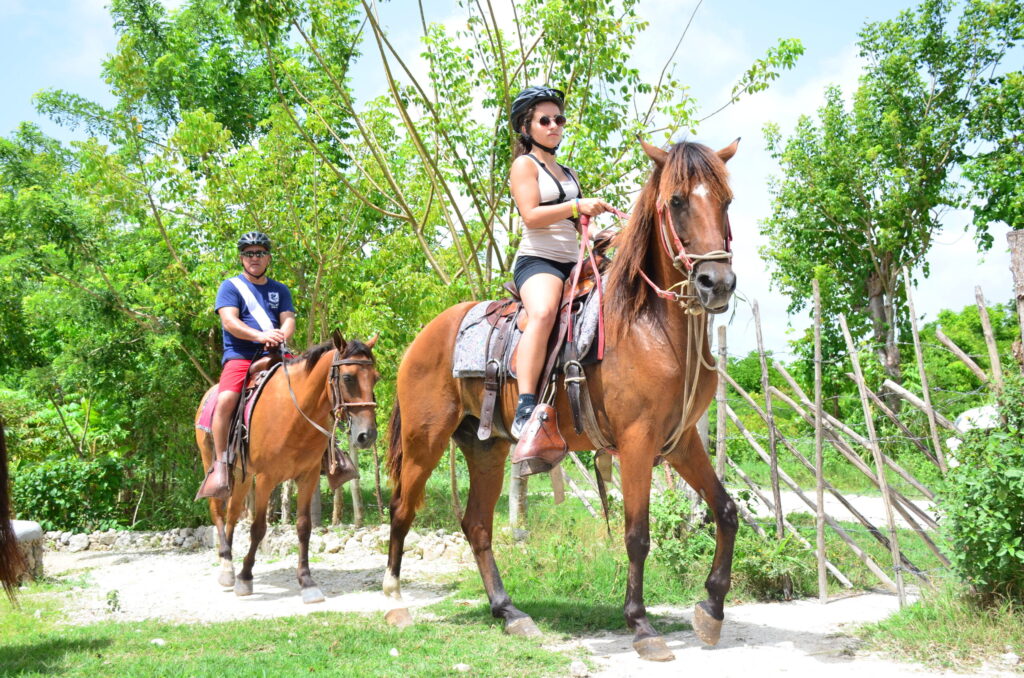 Two tourists wearing safety helmets horseback riding on a scenic dirt trail surrounded by lush green trees.